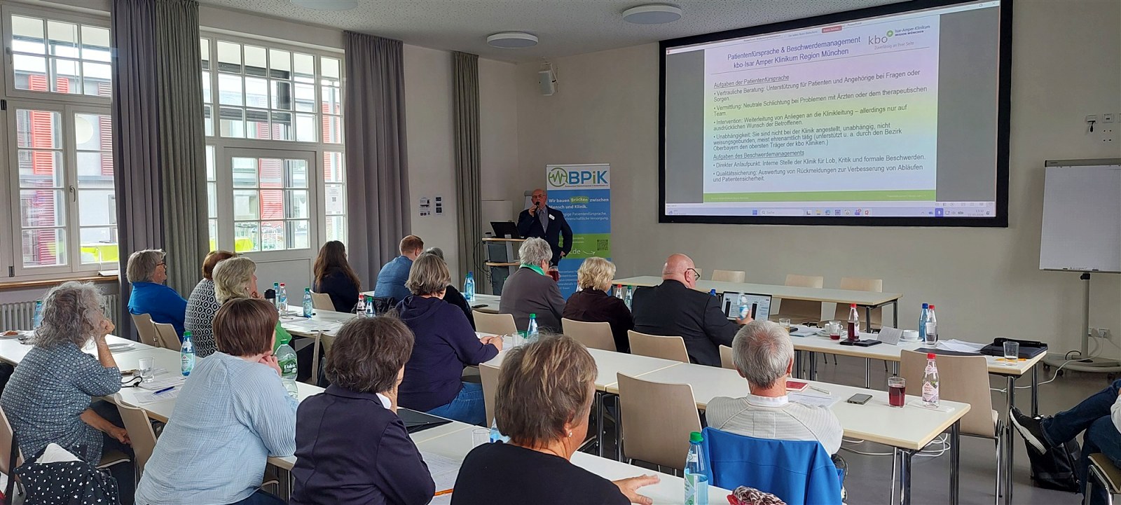 Heinz Wamser am Rednerpult, im Tagungsraum Teilnehmende und Leinwand mit Folie zu Patientenfürsprache und Beschwerdemanagement